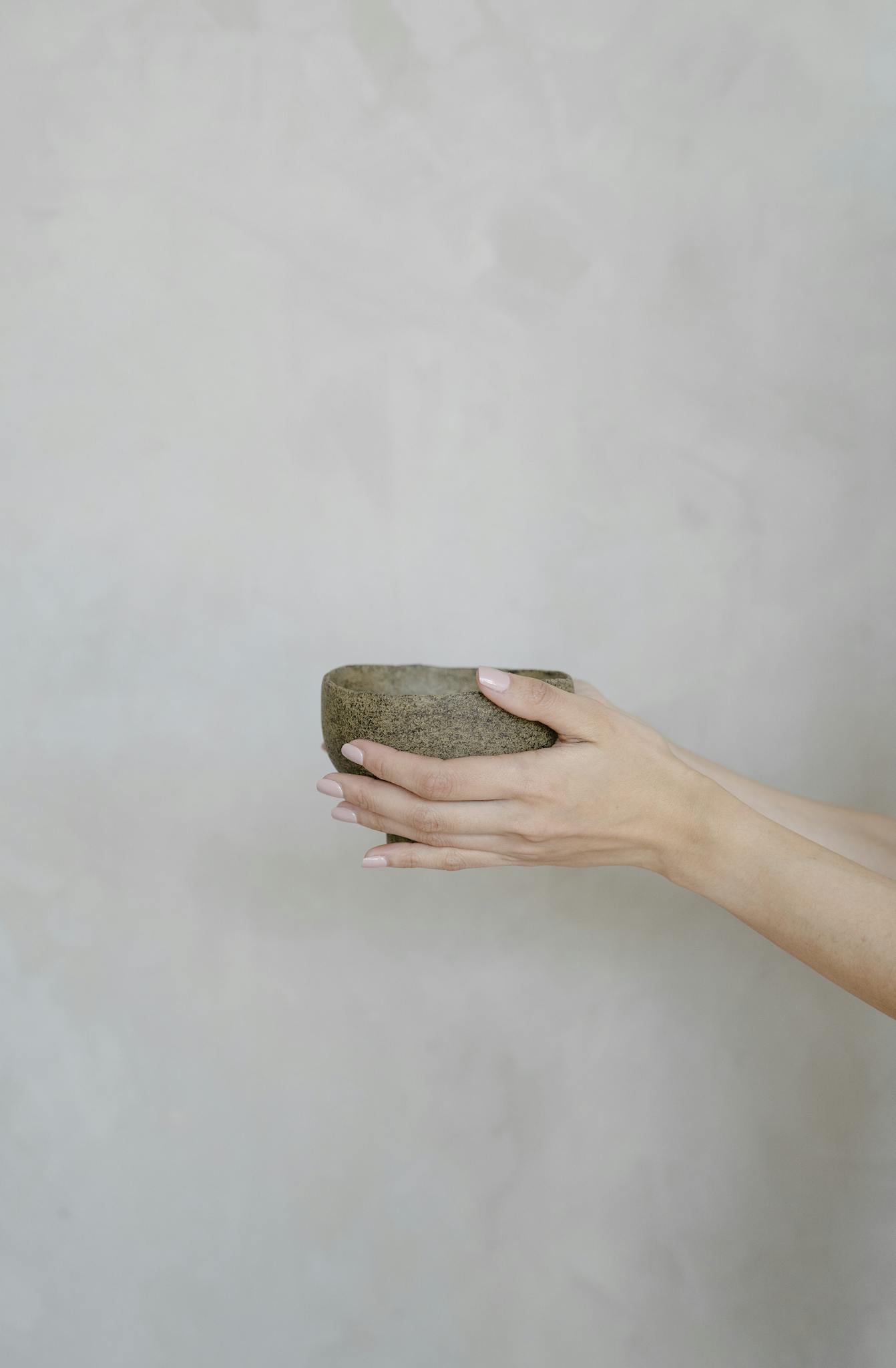 Close-up of hands holding a rustic handmade stoneware bowl against a grey background.
